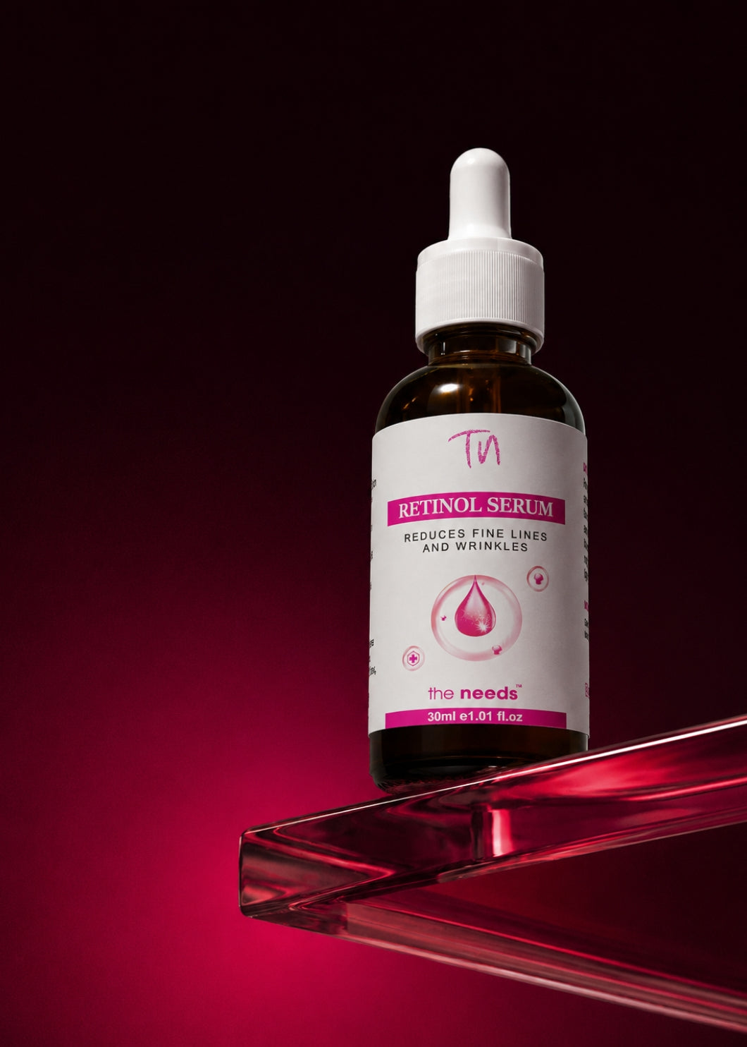 A 30ml amber glass dropper bottle of 'The Needs' Retinol Serum, featuring a white label with pink accents, displayed on a clear acrylic stand against a dark magenta gradient background.