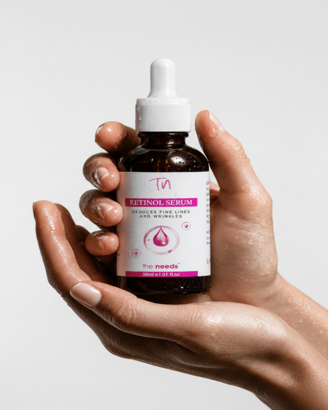A hand holding a small amber dropper bottle of retinol serum against a smooth gray background. The bottle has a white label with pink text and graphics, and the visible brand reads 'the needs'.